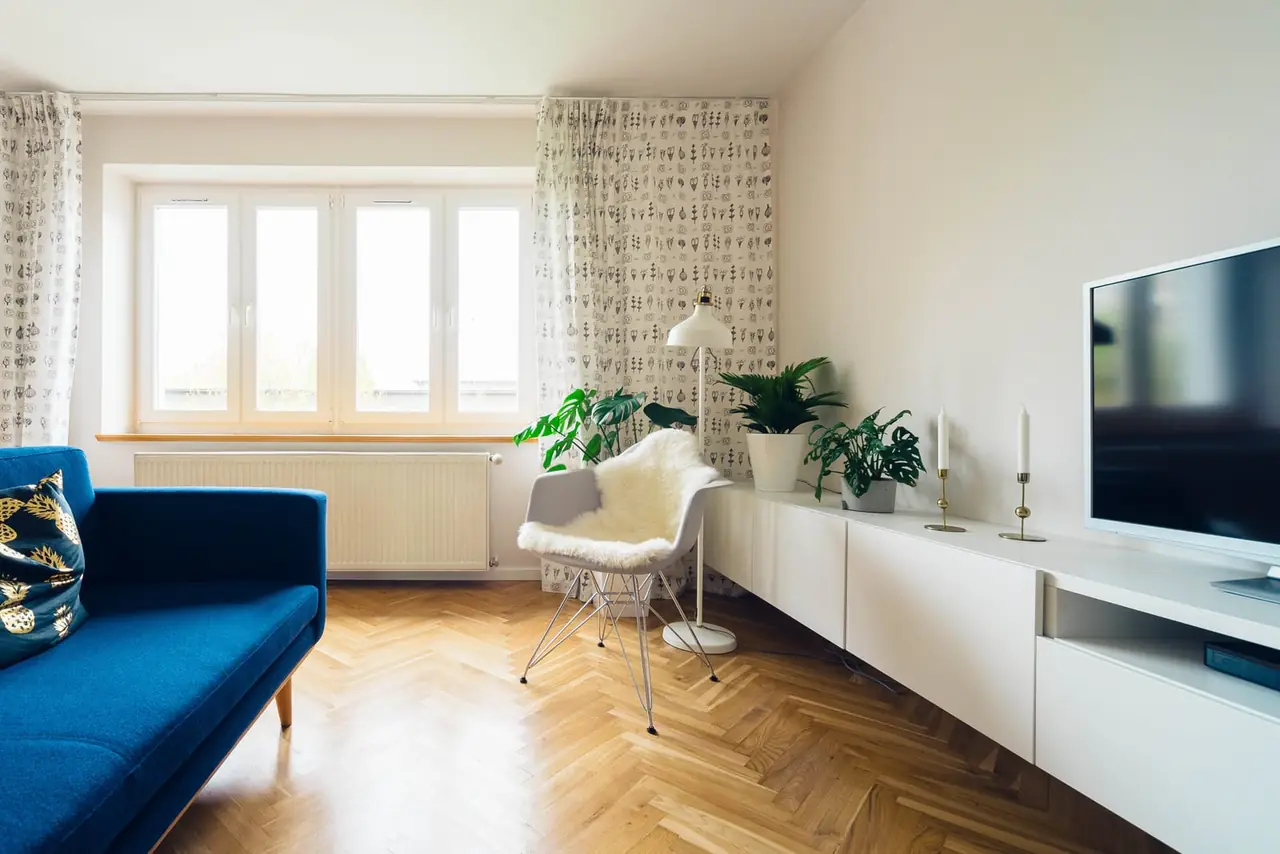 Salon lumineux avec parquet en point de Hongrie, canapé bleu, fauteuil blanc garni d'une peau de mouton et plantes vertes près d'une grande fenêtre