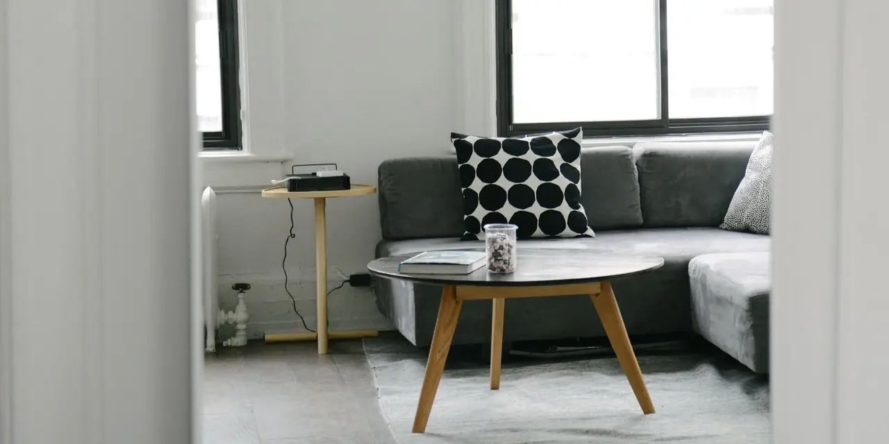 Salon aux murs blancs vu par l'embrasure d'une porte : canapé d'angle gris, coussin à pois noirs, table basse ronde en bois et hautes fenêtres
